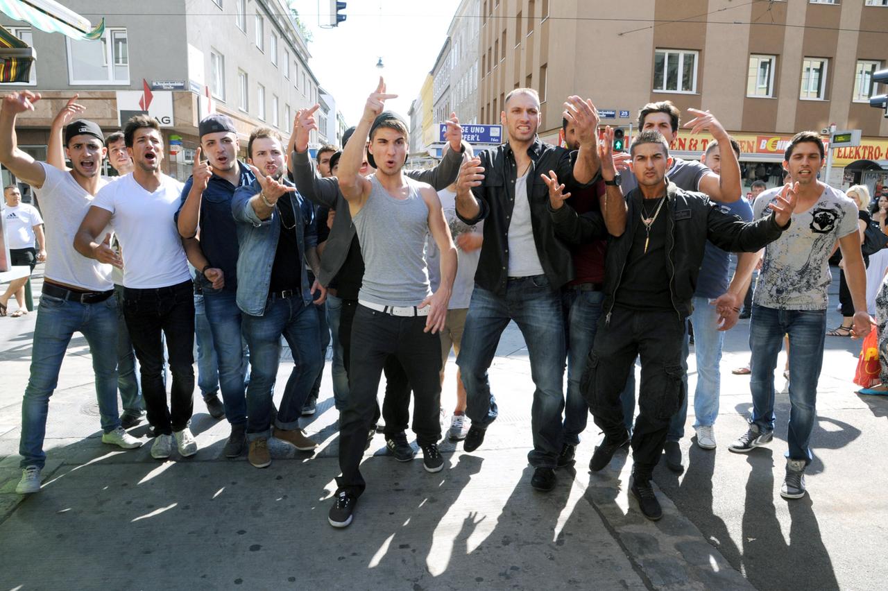 Das Bild zeigt eine Gruppe von mehreren jungen Männern, die auf einer belebten Straße stehen. Sie scheinen aufgeregt und gestikulieren mit ihren Händen, während sie in die Kamera schauen. Die Männer tragen verschiedene Kleidungsstile, darunter T-Shirts, Jeans und Lederjacken. Einige von ihnen haben aufgekrempelte Ärmel oder tragen Sonnenbrillen.   Im Hintergrund sind Gebäude zu sehen, die in verschiedenen Farben gestrichen sind, und es gibt einige Geschäfte, deren Schilder teilweise sichtbar sind. Die Szene wirkt lebhaft und vermittelt eine Stimmung von Spannung. Es ist Tageslicht, und die Schatten der Männer fallen auf den Boden.   Die Umgebung scheint städtisch zu sein, mit einer Mischung aus Wohn- und Geschäftshäusern.