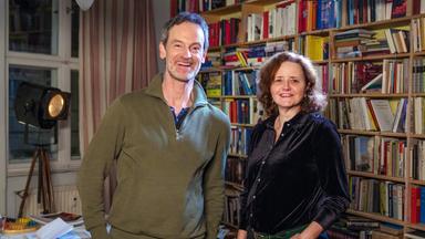 Schauspieler Jörg Hartmann und Schriftstellerin Julia Schoch stehen nebeneinander vor einer Bücherwand. Beide lächeln in die Kamera.