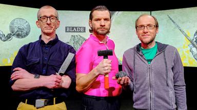 Das Bild zeigt drei Männer auf einer Bühne, die für die TV-Sendung "Science Busters" auftreten. Im Hintergrund sind zwei große, projizierte Leinwände zu sehen, die illustrative Grafiken darstellen.   Der linke Mann hat eine Brille, trägt ein dunkles Hemd und hält ein Schmiedewerkzeug in der Hand. Er wirkt ernst und hat die Arme vor der Brust verschränkt.   Der zentrale Mann trägt ein auffälliges, knallpinker T-Shirt und hält einen Hammer in der rechten Hand. Er schaut in die Kamera und wirkt entschlossen.   Der rechte Mann trägt ein graues Sweatshirt über einem grünen T-Shirt und hält einen kleinen, dunklen Gegenstand in der Hand. Er hat ein freundliches Lächeln auf dem Gesicht.   Die gesamte Szene ist in einem düsteren, aber professionellen Licht gehalten, das die Aufmerksamkeit auf die Darsteller lenkt.