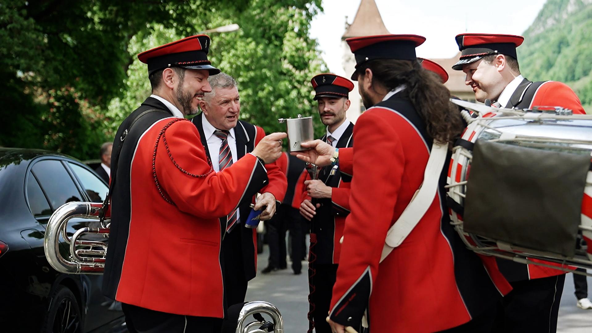 Sechs Männer in rot-schwarzen Jacken mit Musikinstrumenten. Ein Flachmann wird im Zentrum der Gruppe herumgereicht.