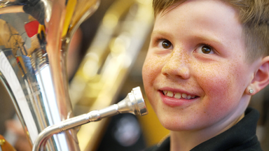 Das Bild zeigt einen Jungen, der ein Musikinstrument, vermutlich eine Tuba, hält. Er hat eine fröhliche Ausstrahlung und lächelt. Der Junge ist möglicherweise zwischen 8 und 12 Jahren alt und hat kurze, blonde Haare. Sein Gesicht ist von Sommersprossen übersät, und er trägt ein schwarzes T-Shirt. Er hält das Mundstück der Tuba in der Nähe seines Gesichts. Im Hintergrund sind unscharfe Silhouetten anderer Musikinstrumente und möglicherweise weiterer Personen zu erkennen, die ebenfalls Musikinstrumente spielen oder vorbereiten. Die Beleuchtung im Raum ist warm und freundlich. Das Bild vermittelt eine Stimmung der Vorfreude und des gemeinschaftlichen Musizierens.