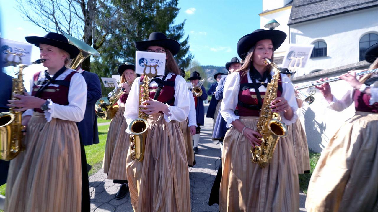 Das Bild zeigt eine Gruppe von Musikern, die in traditioneller Tiroler Tracht auf einem Weg marschieren. Die Musikerinnen tragen lange, hellbraune Röcke und weiße Blusen mit dunklen Westen. Auf ihren Köpfen haben sie schwarze Hüte, die eine breite Krempe besitzen. Die Mehrheit der Musikerinnen hält Saxophone in den Händen, während einige auch andere Blasinstrumente wie Flöten tragen.   Im Hintergrund sind Bäume zu sehen, und es scheint ein sonniger Tag zu sein, da die Sonne hell am Himmel scheint. Hinter den Musikern ist ein Gebäude mit weißen Wänden und einem Dach zu erkennen, das Anzeichen einer traditionellen Architektur aufweist. Die Szenerie vermittelt eine festliche Atmosphäre, die mit der Tiroler Kultur und der Blasmusiktradition verbunden ist.