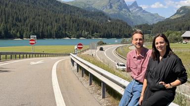 Auf einer kurvigen Straße sitzen ein Mann und eine Frau auf einem Geländer. Im Hintergrund sind Berge und ein See sichtbar. Es ist Tag, der Himmel ist blau mit einigen Wolken.