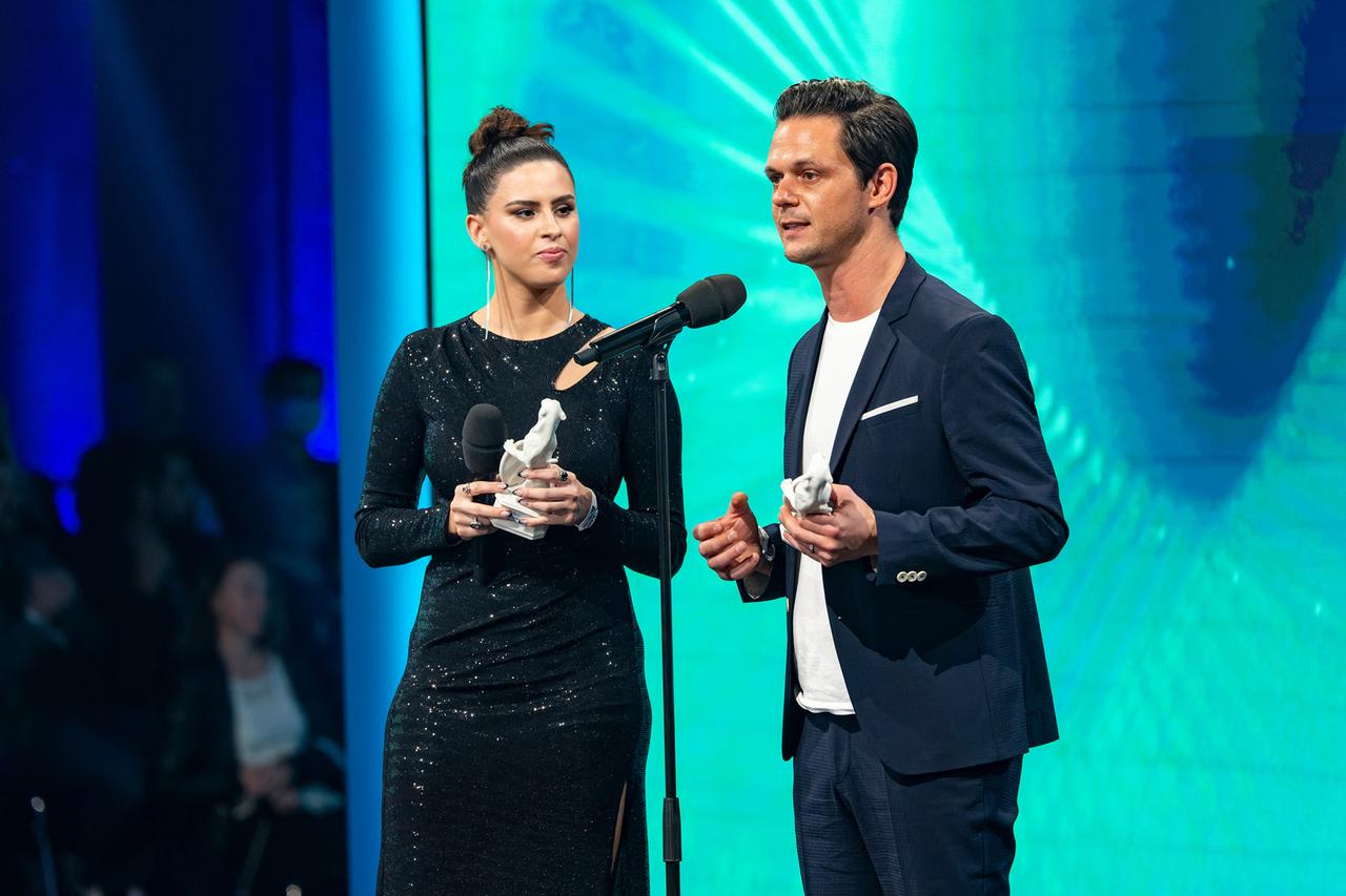 Blauer Panther 2022: Salwa Houmsi und Jo Schück. Frau in schwarzem Abendkleid und Mann im Anzug vor einem türkisfarbenen Hintergrund. 