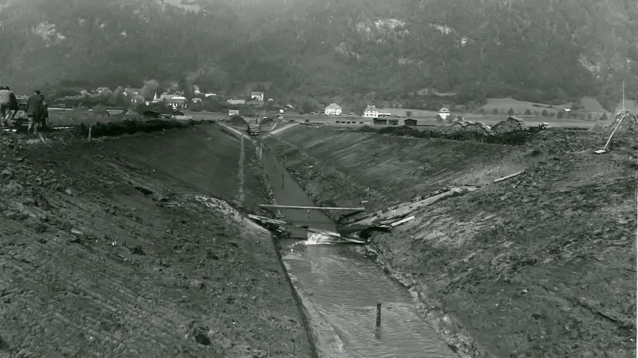 Das Bild zeigt eine historische Szenerie des Bleistätter Moors in Kärnten. Im Vordergrund verläuft ein schmaler Wasserkanal, der durch ein ausgehobenes Gelände von leicht erhöhten Ufern flankiert wird. Auf der linken Seite sind einige Menschen zu sehen, die mit Werkzeugen arbeiten. Im Hintergrund sind mehrere Gebäude, darunter einige Wohnhäuser und landwirtschaftliche Anlagen, erkennbar. Dahinter erhebt sich eine bewaldete Hügelkette.  Die Landschaft ist weitgehend kahl und zeigt deutlich die Spuren der landwirtschaftlichen Nutzung. Der Himmel ist hell und die Wetterbedingungen scheinen klar zu sein. Ingesamt vermittelt das Bild den Eindruck von Arbeit und Veränderung in der Natur, die mit der landwirtschaftlichen Umnutzung des Moores verbunden ist.
