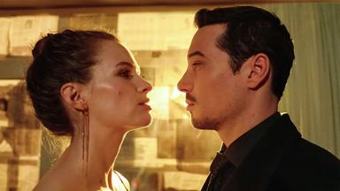 "Blindspot": Anara van Veeren (Marlene Tanczik) und Max van Veeren (Klaus Steinbacher) stehen sich eng gegenüber und schauen sich gegenseitig mit ernster Miene an. Anara hält in ihrer rechten Hand eine brennende Zigarette. Durch das mit Zeitungspapier verdunkelte Fenster scheint gelbes Licht.