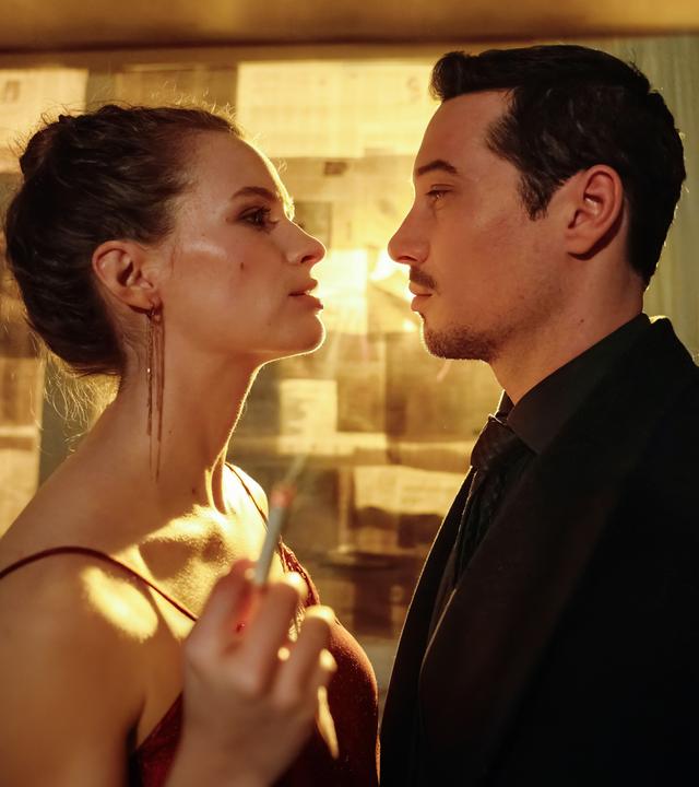 "Blindspot": Anara van Veeren (Marlene Tanczik) und Max van Veeren (Klaus Steinbacher) stehen sich eng gegenüber und schauen sich gegenseitig mit ernster Miene an. Anara hält in ihrer rechten Hand eine brennende Zigarette. Durch das mit Zeitungspapier verdunkelte Fenster scheint gelbes Licht.