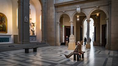 Besicher sitzt in Museumraum auf einer Bank und betrachtet Kunstwerke.