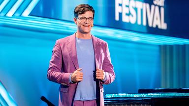 Kabarettist steht mittig auf der Bühne, er lächelt und blickt in Richtung der Kamera. Sein Anzug ist altrosa und er trägt ein hellblaues Shirt. Der Hintergrund ist blau beleuchtet.