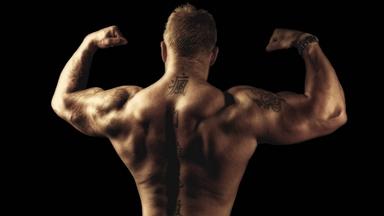 Rückansicht des Oberkörpers eines Bodybuilders mit Tatoos, der mit erhobenen Armen seine Muskeln spielen lässt.