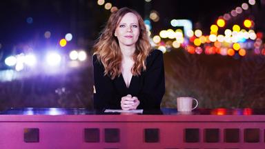 Das Bild zeigt Sarah Bosetti sitzend an einem langen Pult.