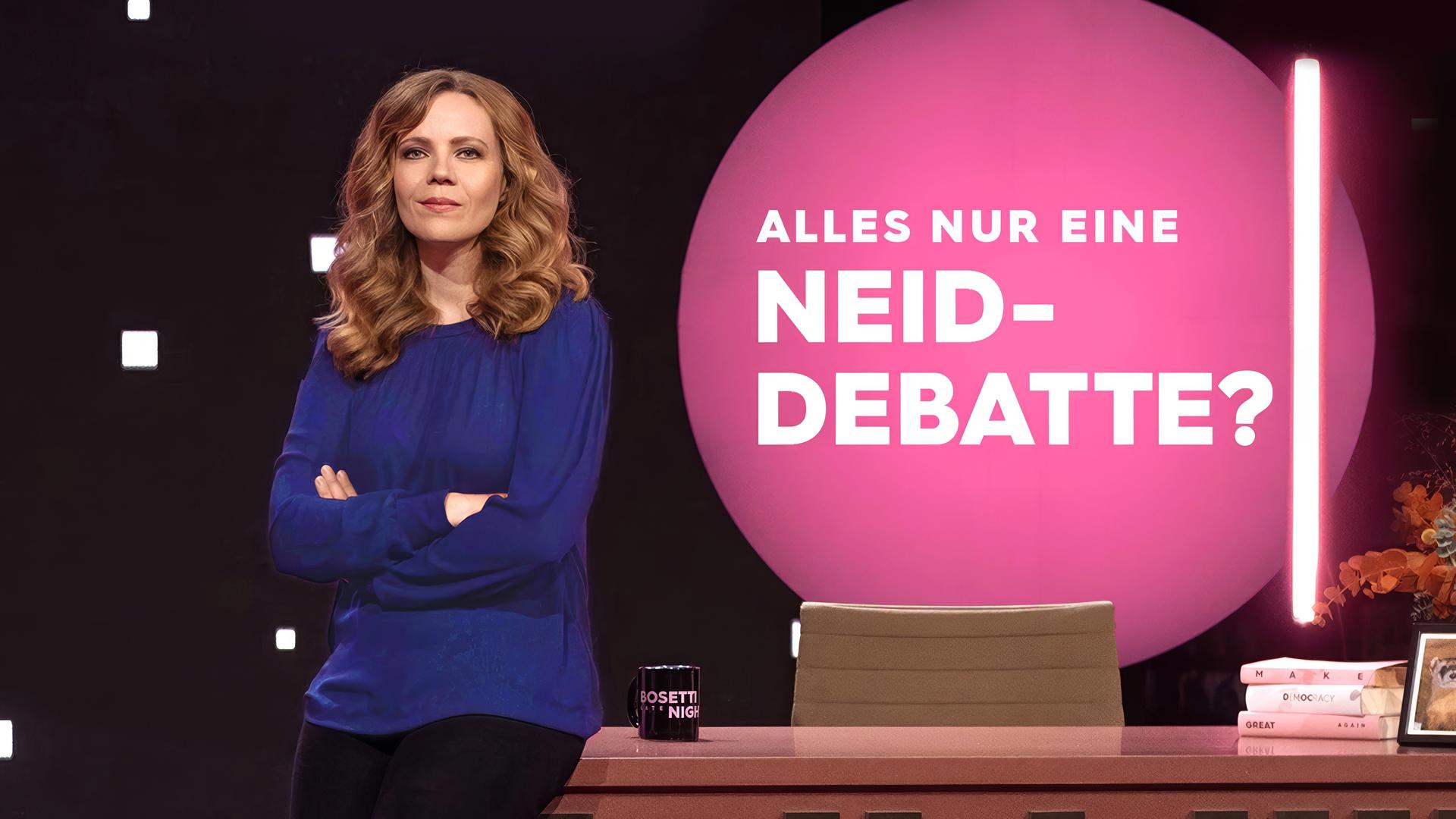 Sarah Bosetti steht auf der linken Seite des Bildes. Sie trägt ein dunkelblaues Oberteil und lehnt mit der Hüfte an einem Schreibtisch. Auf der linken Seite des Tisches steht eine Tasse, rechts liegen drei Bücher übereinande