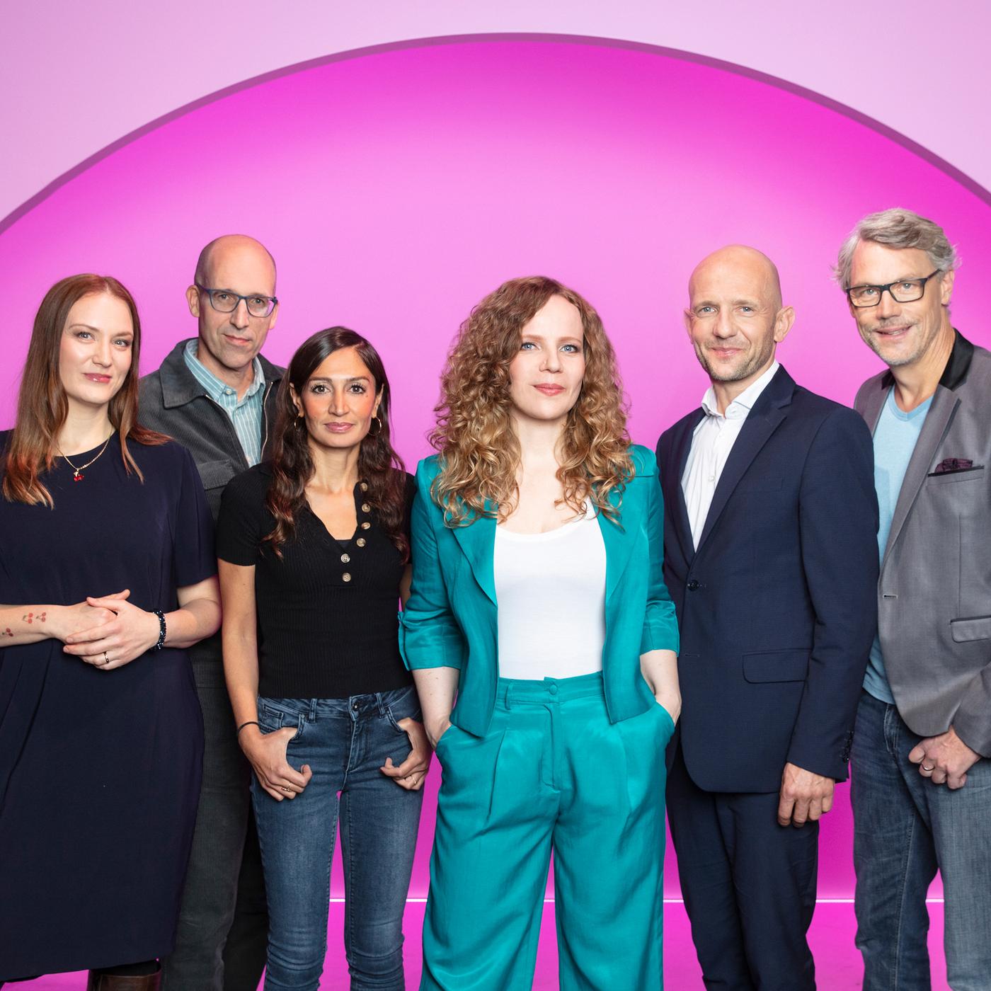 Sarah Bosetti steht in der Mitte des Bildes. Auf ihrer linken Seite stehen drei Personen, auf der rechten zwei weitere. Der Hintergrund ist rosa. Sarah Bosetti trägt einen Zweiteiler in türkis.