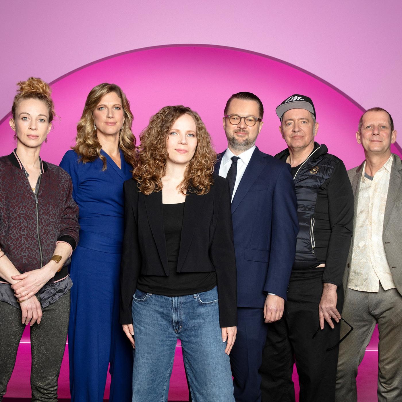 Sarah Bosetti steht in der Mitte des Bildes. Der Hintergrund ist rosa. Es sind 6 Personen abgebildet, zwei links von Sarah Bosetti und drei auf ihrer rechten Seite. Sarah Bosetti trägt eine Jeans, ein schwarzes Oberteil sowie einen kurzen schwarzen Blazer.