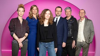 Sarah Bosetti steht in der Mitte des Bildes. Der Hintergrund ist rosa. Es sind 6 Personen abgebildet, zwei links von Sarah Bosetti und drei auf ihrer rechten Seite. Sarah Bosetti trägt eine Jeans, ein schwarzes Oberteil sowie einen kurzen schwarzen Blazer.