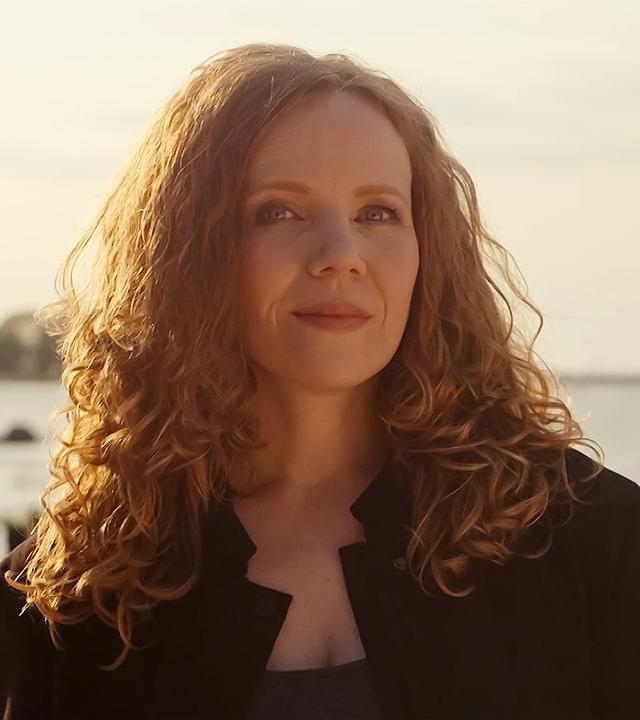 Sarah Bosetti steht in der Mitte des Bildes. Im Hintergrund ist ein Sonnenuntergang und Wasser, das Licht ist hell. Sarah Bosetti trägt eine schwarze Jacke, ihre Haare sind offen. 