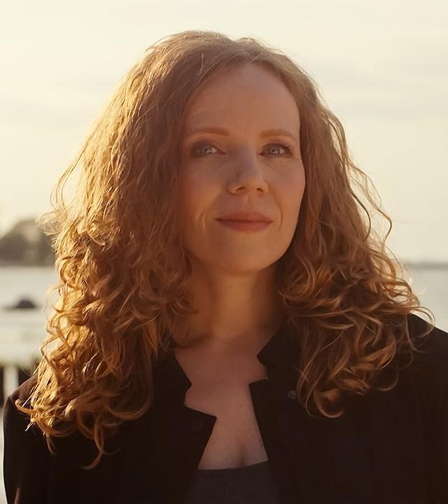 Sarah Bosetti steht in der Mitte des Bildes. Im Hintergrund ist ein Sonnenuntergang und Wasser, das Licht ist hell. Sarah Bosetti trägt eine schwarze Jacke, ihre Haare sind offen. 