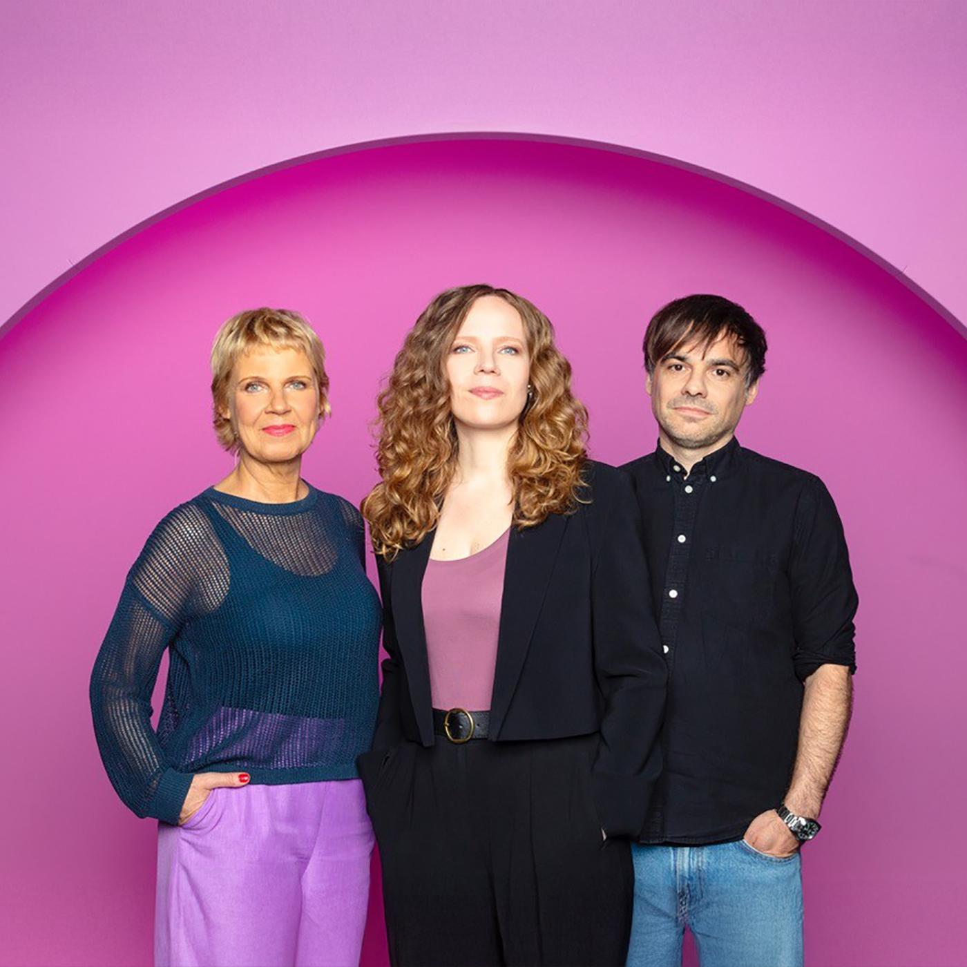 Sarah Bosetti steht in der Mitte des Bildes. Links von ihr steht Cordula Stratmann und rechts von Sarah Bosetti ist Sebastian Lehmann. Der Hintergrund ist rosa.