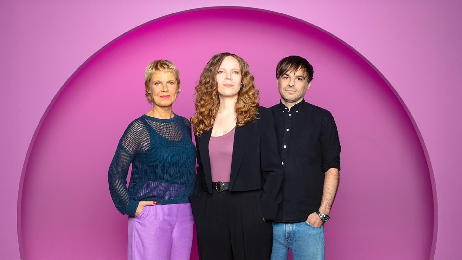 Sarah Bosetti steht in der Mitte des Bildes. Links von ihr steht Cordula Stratmann und rechts von Sarah Bosetti ist Sebastian Lehmann. Der Hintergrund ist rosa.