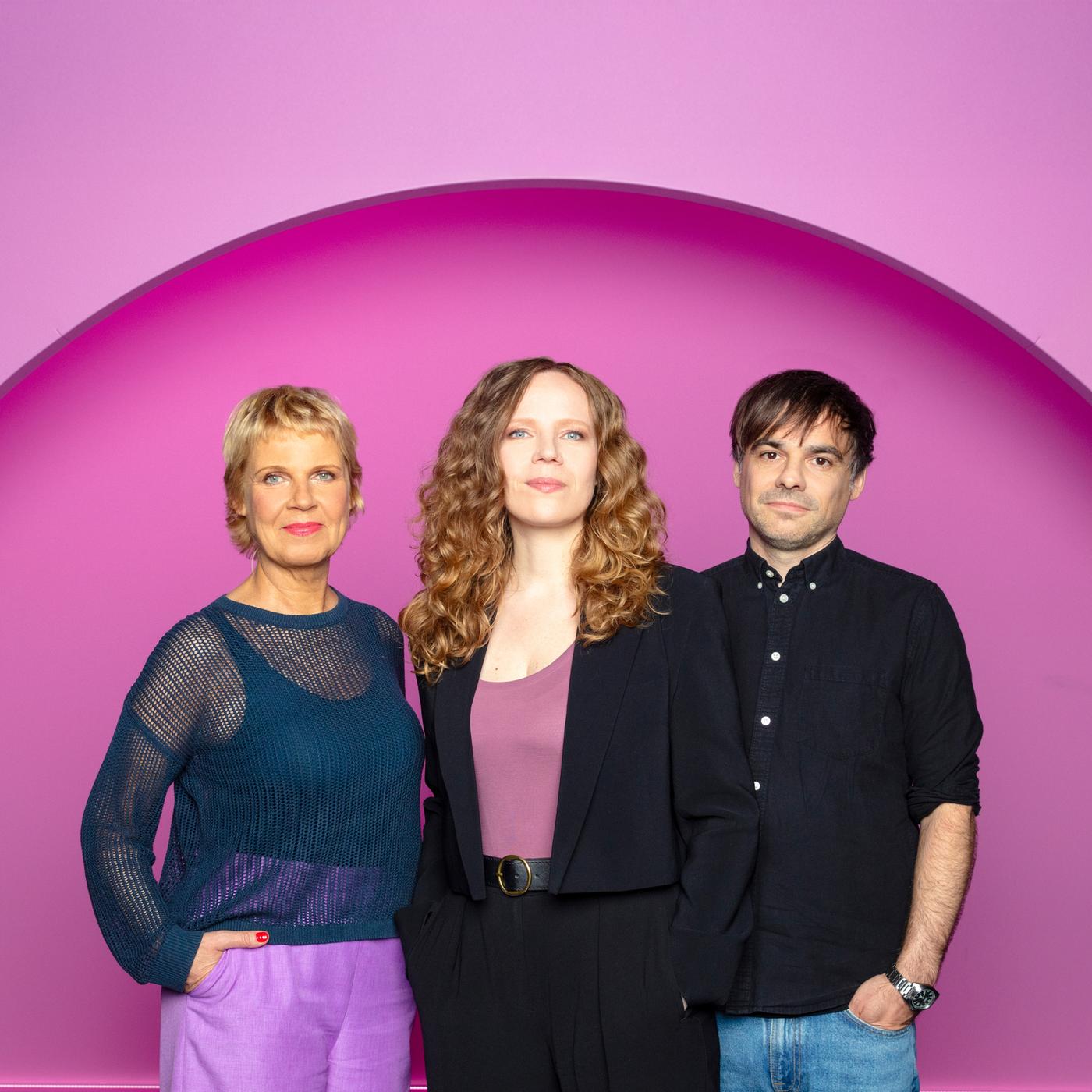 Sarah Bosetti steht in der Mitte des Bildes. Links von ihr steht Cordula Stratmann und rechts von Sarah Bosetti ist Sebastian Lehmann. Der Hintergrund ist rosa.