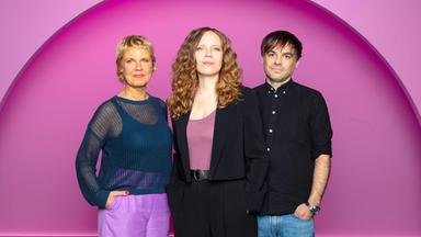 Sarah Bosetti steht in der Mitte des Bildes. Links von ihr steht Cordula Stratmann und rechts von Sarah Bosetti ist Sebastian Lehmann. Der Hintergrund ist rosa.