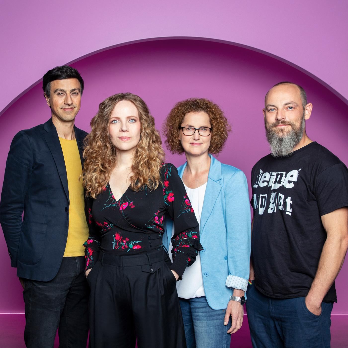 Auf dem Bild stehen vier Personen nebeneinander. Sarah Bosetti ist die zweite Person von Link.  Sie trägt ihr Haar offen und ist schwarz gekleidet. Der Hintergrund ist rosa.