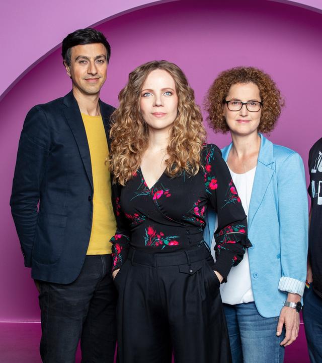 Auf dem Bild stehen vier Personen nebeneinander. Sarah Bosetti ist die zweite Person von Link.  Sie trägt ihr Haar offen und ist schwarz gekleidet. Der Hintergrund ist rosa.