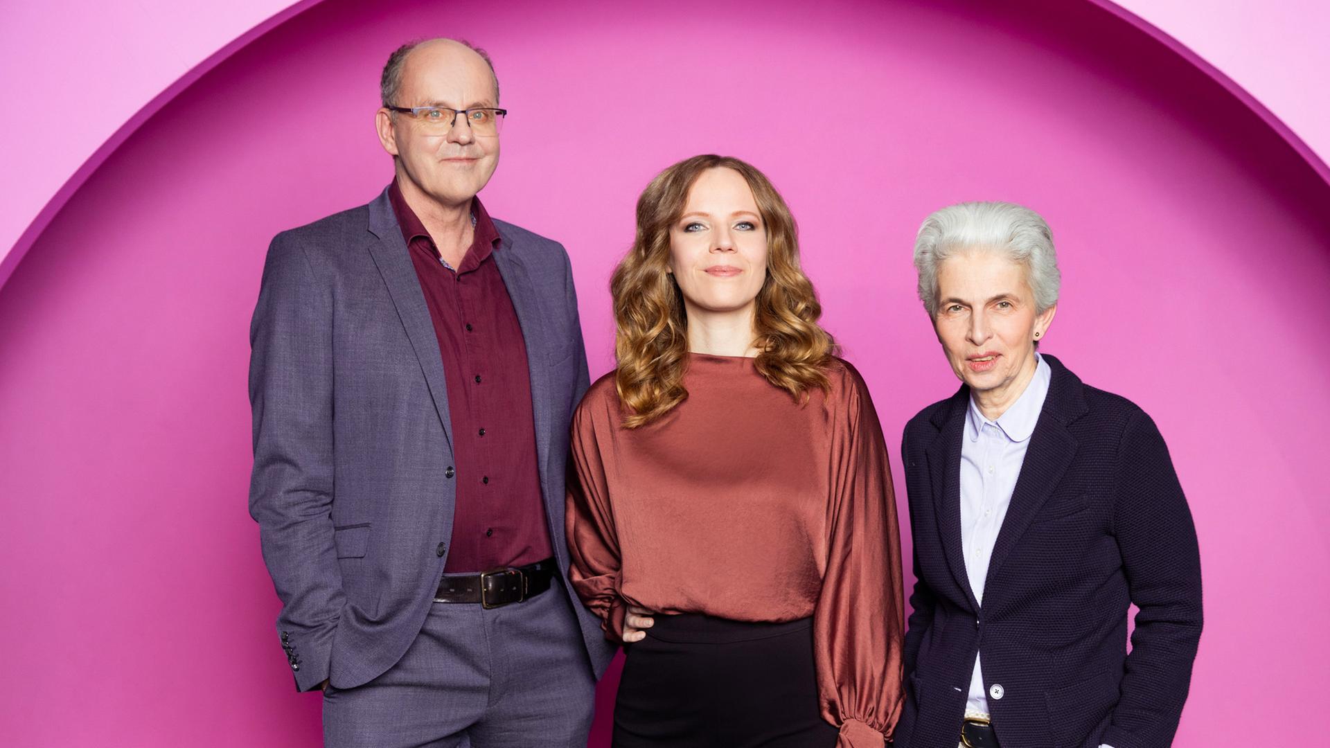 Auf dem Bild ist Sarah Bosetti mittig zu sehen. Links neben ihr steht Andreas Hasenclever. Auf der rechten Seite des Bildes ist Marie-Agnes Strack-Zimmermann. Der Hintergrund ist rosa.