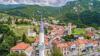 Panoramaaufnahme von Travnik mit Moschee, Minaretten, Uhrturm, roten Dächern und umliegenden grünen Bergen.