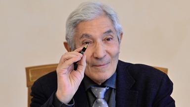 Porträtfoto eines Mannes mit zurückgebundenen grauen Haaren und runder Metallbrille, der in die Kamera lächelt.