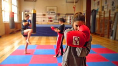 Eine Frau in Boxposition mit roten Boxhandschuhen auf denen ein Frauenpower Symbol zu sehen ist.