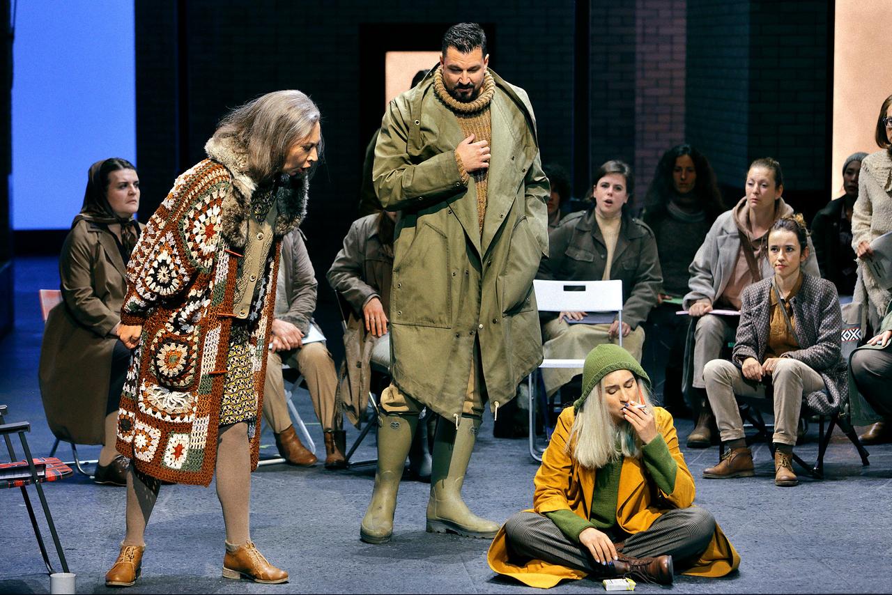 Mehrere Personen auf einer Theaterbühne. Eine Frau im gelben Mantel sitzt auf dem Boden und raucht.