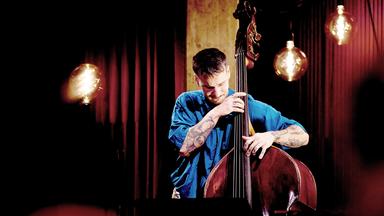 Ein Mann in blauem Shirt und tätowierten Unterarmen spielt einen Kontrabass. Im Hintergrund sind runde Hängeleuchten und ein roter Vorhang.