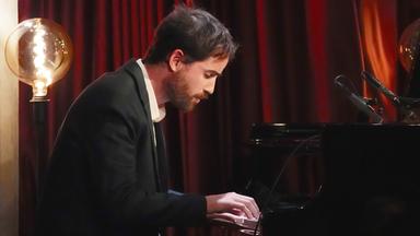 Ein Mann in dunklem Anzug sitzt gebeugt über die Klaviatur an einem Klavierflügel. Hinter ihm steht eine Standleuchte. Im Hintergrund ist ein roter Vorhang zu sehen.