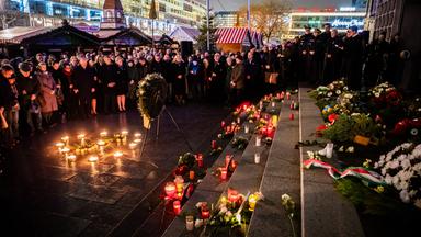 Eine Menschenmenge steht bei nacht um auf dem Boden aufgestellte Kerzen und einem Kranz vor einer Treppe, auf der noch mehr Votivkerzen stehen. Im Hintergrund ist ein Weihnachtsmarkt zu sehen.