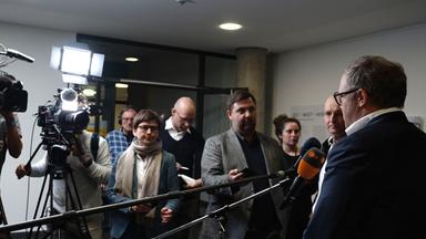 Rückansicht eines Mannes, der vor einer Gruppe Journalisten mit Kameras und Mikrophonen steht.