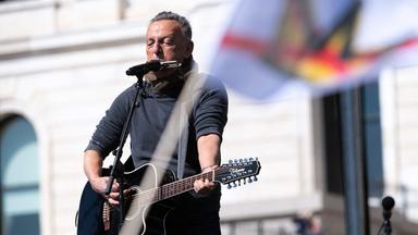 Mann spielt Gitarre und Mundharmonika, im Vordergrund Fahne mit durchgestrichener Krone.