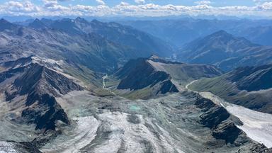 Das Bild zeigt eine atemberaubende Berglandschaft in den Alpen. Im Vordergrund sind große, graue Gletscherflächen sichtbar, die sich in Wellen über das Terrain ausbreiten. Diese Gletscher zeigen deutliche Zeichen des Rückgangs, mit großflächigen, schimmernden Eisschichten, die sich von den Felsen abheben.  Die Umgebung ist geprägt von zerklüfteten Berghängen und schroffen Gipfeln, die in der Ferne in sanfteren grünen Hügeln übergehen. Mehrere scharfe, dunkle Felskanten sind im Vordergrund zu sehen, während die Höhenlagen hinter den Gletschern sich in sanften Blautönen und Wolken verlieren. Der Himmel ist klar, und einige Wolken schweben über den Bergen. Eine Wasserstraße schlängelt sich durch die Landschaft, was auf einen möglichen Fluss oder Bach hindeutet.  Diese Szene vermittelt eine eindrückliche Vorstellung von der Schönheit der Natur und der fragilen Situation in den Alpen, insbesondere in Bezug auf den Klimawandel.