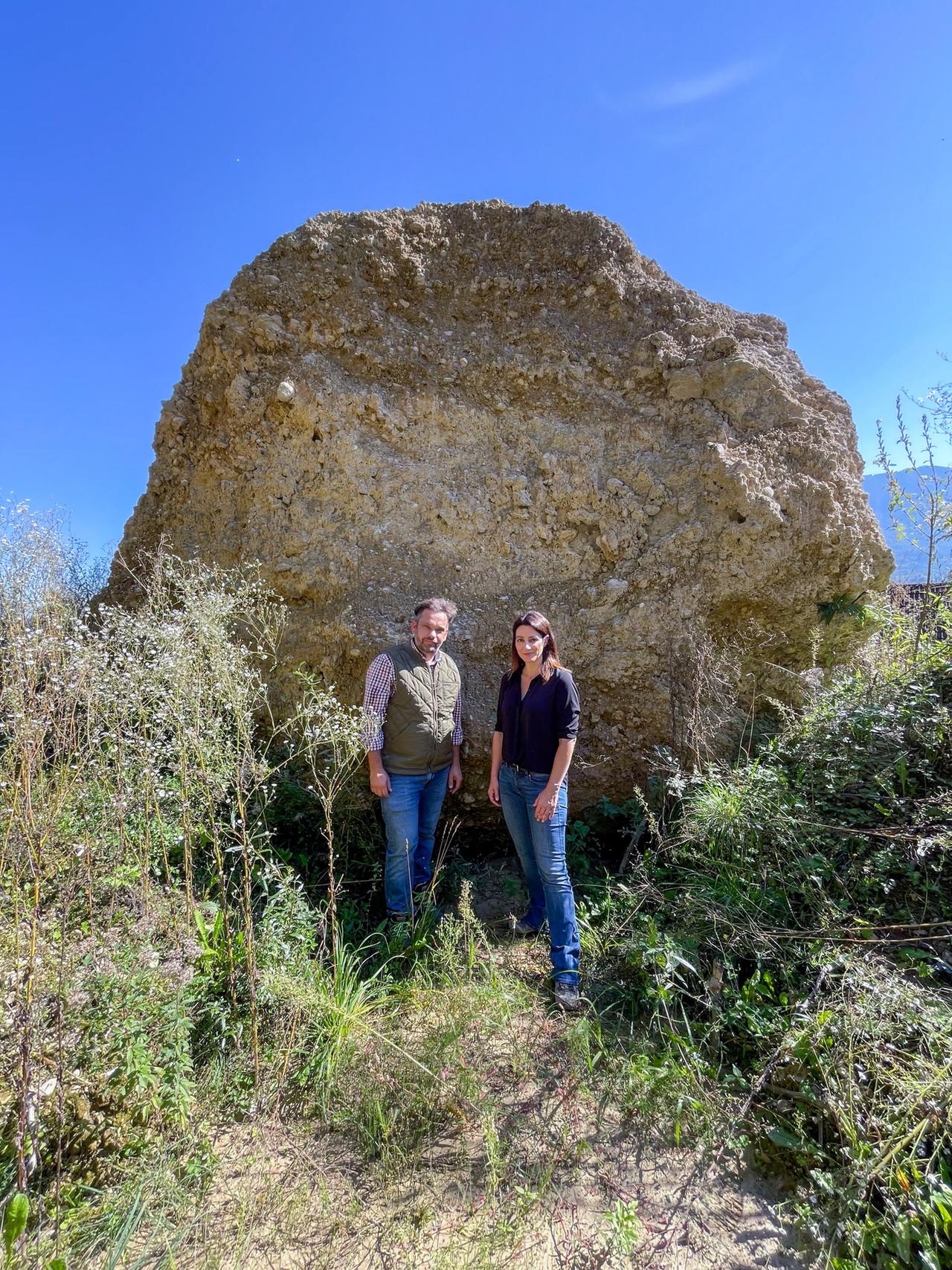 Das Bild zeigt zwei Personen, die vor einem großen Felsen stehen. Die Person links trägt eine Weste und ein kariertes Hemd, während die Person rechts ein schwarzes Oberteil und Jeans trägt. Sie stehen auf grünem, leicht bewachsenem Boden, umgeben von einigen Pflanzen und Gräsern. Im Hintergrund ist ein strahlend blauer Himmel zu sehen, was auf gutes Wetter hinweist. Der Felsen, vor dem sie stehen, ist von der strukturierten Oberfläche schwer und massiv. Das Bild vermittelt eine Atmosphäre, die sowohl die Natur als auch die Herausforderungen des Klimawandels thematisiert.