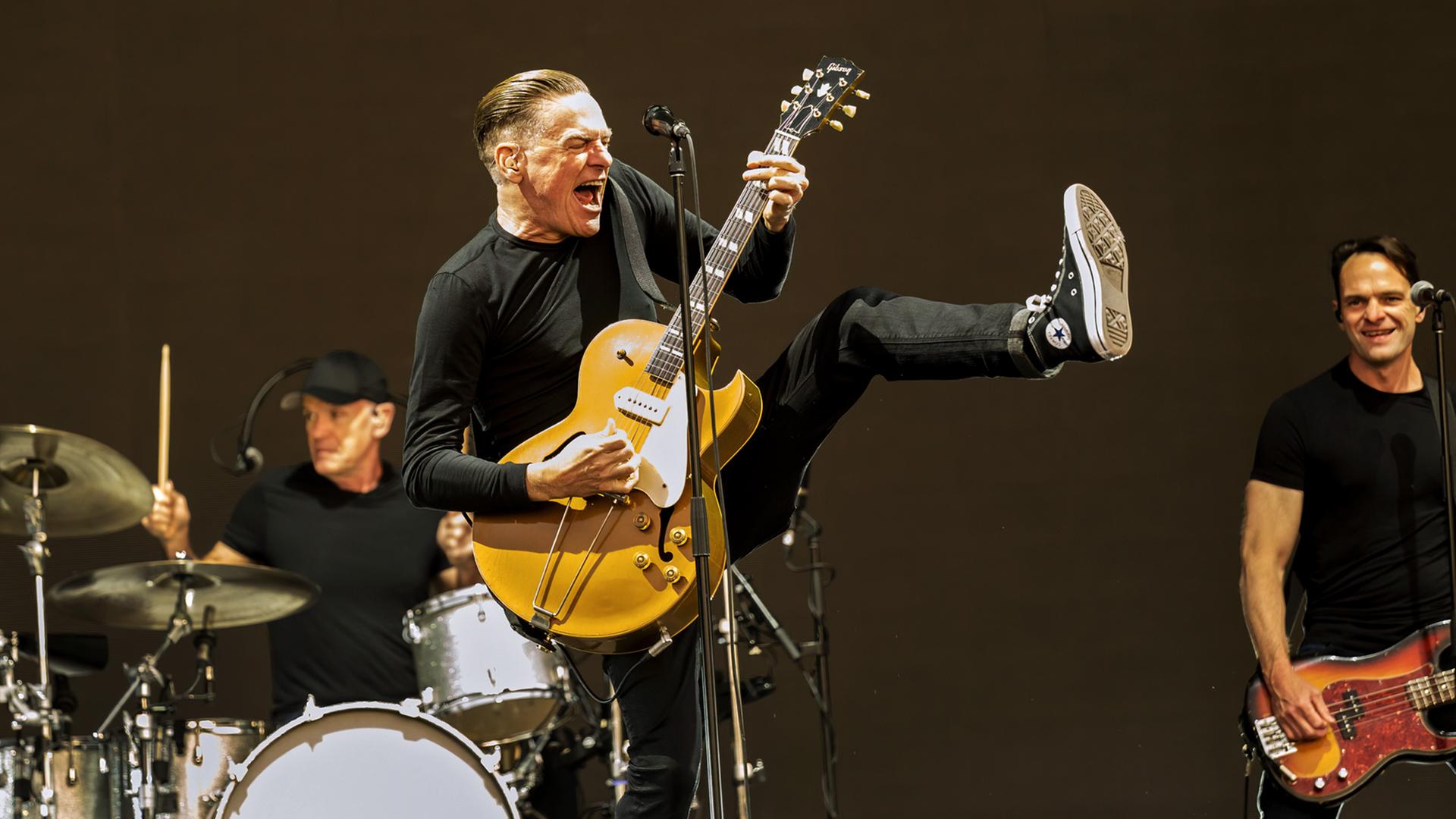 Bryan Adams singt mit seiner Gitarre auf der Bühne. Hinter ihm ist seine Band zu sehen.