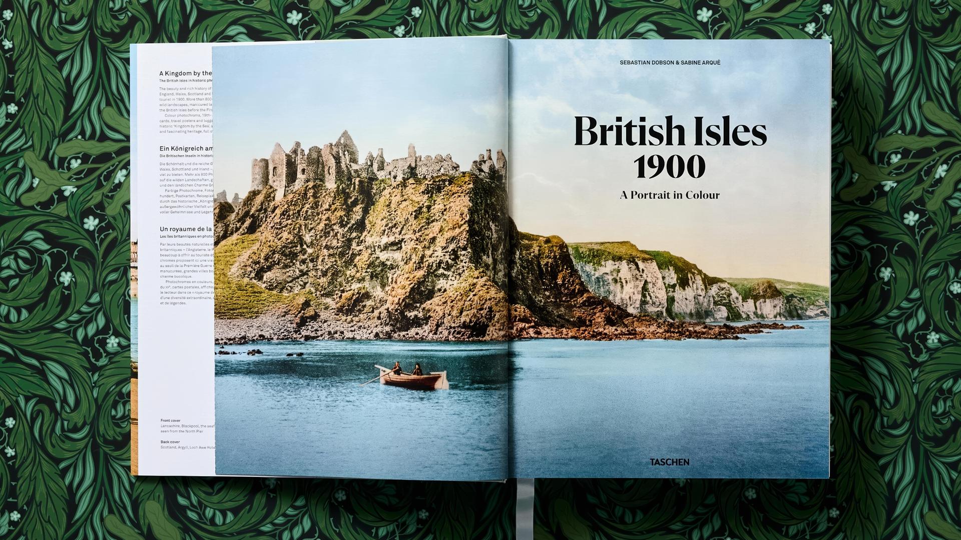 Ein großformatiges Buch liegt aufgeschlagen auf einem mit grünen Blättern bemalten Untergrund. Auf den aufgeschlagenen Seiten ist ein zweiseitiges coloriertes historisches Foto von einer Burgruine auf einer Steilküste zu sehen. Im Vordergrund ist ein Ruderboot auf einem See oder Meer unterwegs. Daneben steht der Titel British Isles 1900