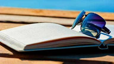 Ein aufgeschlagenes Buch mit einer daraufliegenden Sonnenbrille liegt auf einer Holzterrasse. Im Hintergrund ist das blaue Meer zu sehen.
