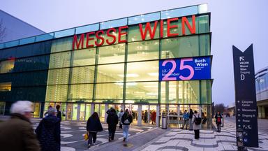 Menschen laufen über einen Platz in einen großen modernen Glasbau, auf dem Messe Wien steht und ein Plakat der Buch Wien 25 hängt.