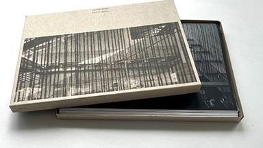 In einer Kassette, die mit dem Foto eines modernen Gebäudes mit viel Glas bedruckt ist, liegt ein Fotobuch.