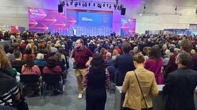 Das Bild zeigt eine große Menschenmenge in einem Innenraum, der vermutlich eine Messe oder ein Festival beherbergt, mit dem Titel "Buch Wien" prominent an einer Wand. Die Bühne ist beleuchtet und mit einem Bildschirm ausgestattet, auf dem Informationen angezeigt werden. Im Vordergrund sind sowohl Männer als auch Frauen zu sehen, die sich in einer wartenden Position befinden; einige sind fotografierend, während andere in Gespräche vertieft sind.  Die Zuhörer sitzen auf schwarzen Stühlen und zeigen eine Vielzahl von Haarfarben und -stilen, sowie unterschiedliche Kleidungsstücke. Die Atmosphäre wirkt lebhaft und erwartungsvoll, da viele Personen auf die Bühne blicken oder sich auf das Geschehen vorbereiten. Die Wände des Raumes sind schlicht und hell, während die beleuchteten Elemente in kräftigen Farben gestaltet sind, die eine ansprechende Umgebung schaffen.