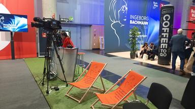 Das Bild zeigt einen Stand auf einer Veranstaltung, vermutlich einer Buchmesse.   Im Vordergrund stehen zwei orangefarbene Liegestühle mit einem Druck, der einen Text und eine Skizze zeigt. Hinter den Stühlen befindet sich ein grün gefärbter Teppich und ein Tisch, an dem eine Person sitzt, die ein Mikrofon und technische Geräte bedient.   Links von dem Tisch steht eine Kamera auf einem Stativ, die auf die Szene gerichtet ist. Im Hintergrund hängt ein großes, hellblaues Banner mit dem Namen „INGEBORG BACHMANN“ und den Lebensdaten 1926-1973-2026. Daneben sind weitere Stände zu sehen, unter anderem mit der Aufschrift „Coffee to go“ auf einem großen Behälter.  Die Umgebung ist hell erleuchtet, und es sind weitere Veranstaltungen oder Stände im Hintergrund sichtbar.