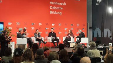 Das Bild zeigt eine Veranstaltungsbühne, die mit einem orangefarbenen Hintergrund dekoriert ist, auf dem die Wörter "Buch Wien", "Inspiration", "Austausch", "Debatte" und "Bildung" stehen. In der Mitte sitzen fünf Personen auf weißen, modernen Stühlen.   Die erste Person auf der linken Seite hält eine Kamera und ist als Fotograf erkennbar. Neben ihm sitzt ein Mann mit einem Hut, der einen grau-gestreiften Pullover trägt. Rechts von ihm ist eine Frau mit langen, dunklen Haaren und einer schwarzhäutigen Bluse mit weißem Muster. In der Mitte sitzt ein Mann in schwarzer Kleidung, der ein Blatt Papier oder ein Notizbuch in der Hand hält und in die Runde blickt. Neben ihm sitzt eine Frau in einem glänzenden, burgunderfarbenen Oberteil und einer schwarzen Hose. Die letzte Person ist ein älterer Mann in schwarzem Outfit, der interessiert zuhört.   Vor der Bühne sind Zuschauer zu sehen, die auf die Diskussion achten, einige klatschen. In der Ecke des Raumes befindet sich ein Lichtstrahler, der die Szene beleuchtet.