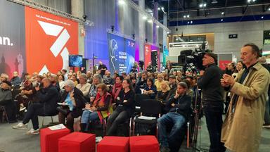 Das Bild zeigt ein Publikum in einem großen Veranstaltungsraum. Im Vordergrund sind mehrere Personen in Sitzposition zu sehen, die auf eine Bühne oder einen Vortrag schauen. Die Zuschauer sind vielfältig, sowohl in Geschlecht als auch in Alter. Einige Personen haben Notizen oder Getränke in der Hand.  Im Hintergrund sind große Banner mit verschiedenen Aufschriften und Logos sichtbar, darunter eines für die Ingeborg-Bachmann-Preise, das in dunkler Schrift auf hellem Grund steht. Weiterhin sieht man Kamerateams und Fotografen, die die Veranstaltung dokumentieren.   Der Boden ist mit einem grauen Teppich bedeckt, und es sind mehrere rote Sitzwürfel im Raum verteilt. Die Raumbeleuchtung ist hell und es sind hohe Decken mit sichtbaren Trägern. Alles in allem vermittelt das Bild eine Atmosphäre der Aufmerksamkeit und des Interesses an der Veranstaltung.
