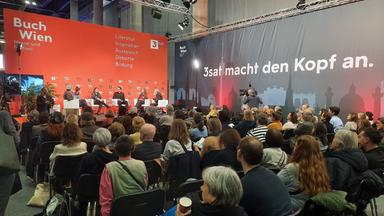 Das Bild zeigt eine Veranstaltung während der Buchmesse "Buch Wien". Im Vordergrund befinden sich mehrere Personen auf einer Bühne, die in weißen Sesseln sitzen. Diese Personen scheinen an einer Diskussion oder einem Panel teilzunehmen. Hinter ihnen steht eine große, rote Wand mit dem Schriftzug "Buch Wien" sowie anderen Texten in weißer Schrift, die Themen wie Literatur, Inspiration und Bildung ansprechen.  Im Publikum sitzen zahlreiche Menschen, die auf die Bühne schauen. Die Zuschauer sind auf schwarzen Stühlen und scheinen interessiert zuzuhören. Einige haben Notizen oder Getränke in den Händen. Im Hintergrund ist eine Kamera mit einem Kameramann positioniert, der die Veranstaltung aufnimmt. Die Atmosphäre wirkt lebhaft und engagiert, typisch für eine literarische Veranstaltung oder ein Festival.