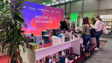 Das Bild zeigt einen Stand auf einer Buchmesse oder ähnlichen Veranstaltung. Im Hintergrund ist ein großes, farbenfrohes Banner mit der Aufschrift „HOUSE OF ROMANCE“ zu sehen, das in Pink- und Lila-Tönen gestaltet ist. Vor dem Banner befindet sich ein Tisch, der mit einer Vielzahl von Bücherstapeln bedeckt ist. Die Titel sind unterschiedlich, einige bieten bunte Cover.  Auf der linken Seite des Tisches steht eine Pflanze in einem Topf. Vor dem Tisch sind mehrere Personen zu sehen, die die Bücher anschauen und durch Broschüren blättern. Die Kleidung der Personen ist leger, und sie scheinen interessiert an dem Angebot. Im Hintergrund sind weitere Stände und eine grüne Fahne sichtbar, die wahrscheinlich zu einem anderen Aussteller gehört. Der Boden des Standes ist mit grauem Teppich ausgelegt, und es gibt einen roten Teppich vor dem Tisch.